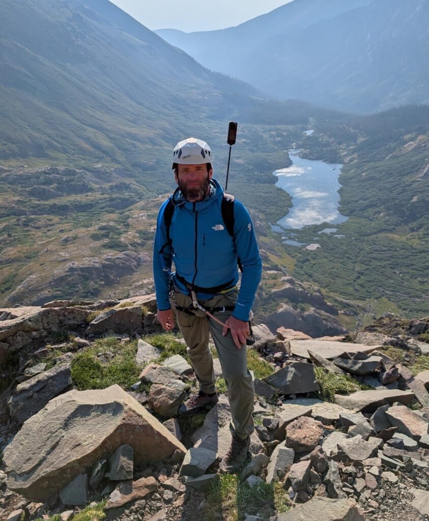 Dima wearing climbing gear above an alpine lake.